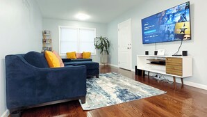 A modern living room with a blue sofa, a coffee table, and a flat-screen TV mounted on the wall. - Modern 3BR 2BA• 5 Min to EWR • 30 Min to NYC  (Elizabeth)