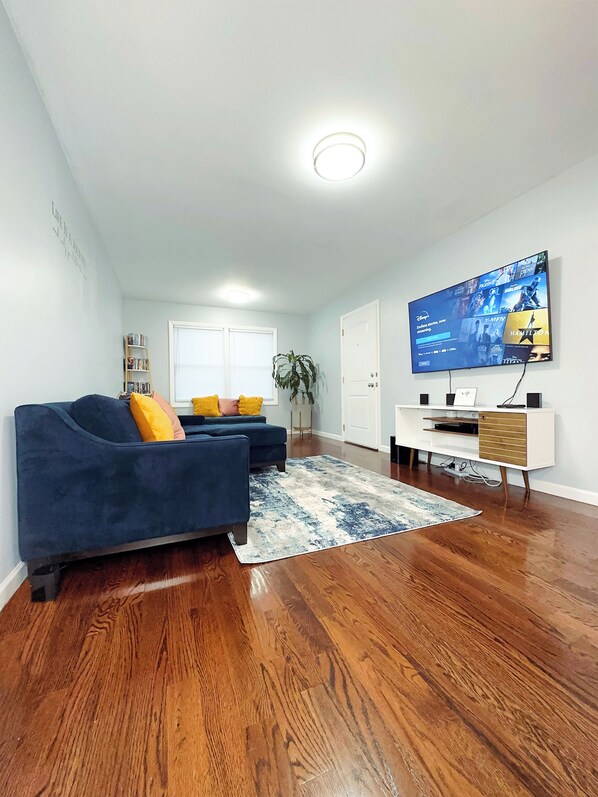 A modern living room with a blue sofa, a coffee table, and a flat-screen TV mounted on the wall. - Modern 3BR 2BA • Near EWR & NYC • Parking (Elizabeth)