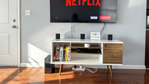 A modern living room with a mounted television displaying the Netflix logo, a white entertainment unit with books and electronic devices, and a wooden floor. - Modern 3BR 2BA• 5 Min to EWR • 30 Min to NYC  (Elizabeth)