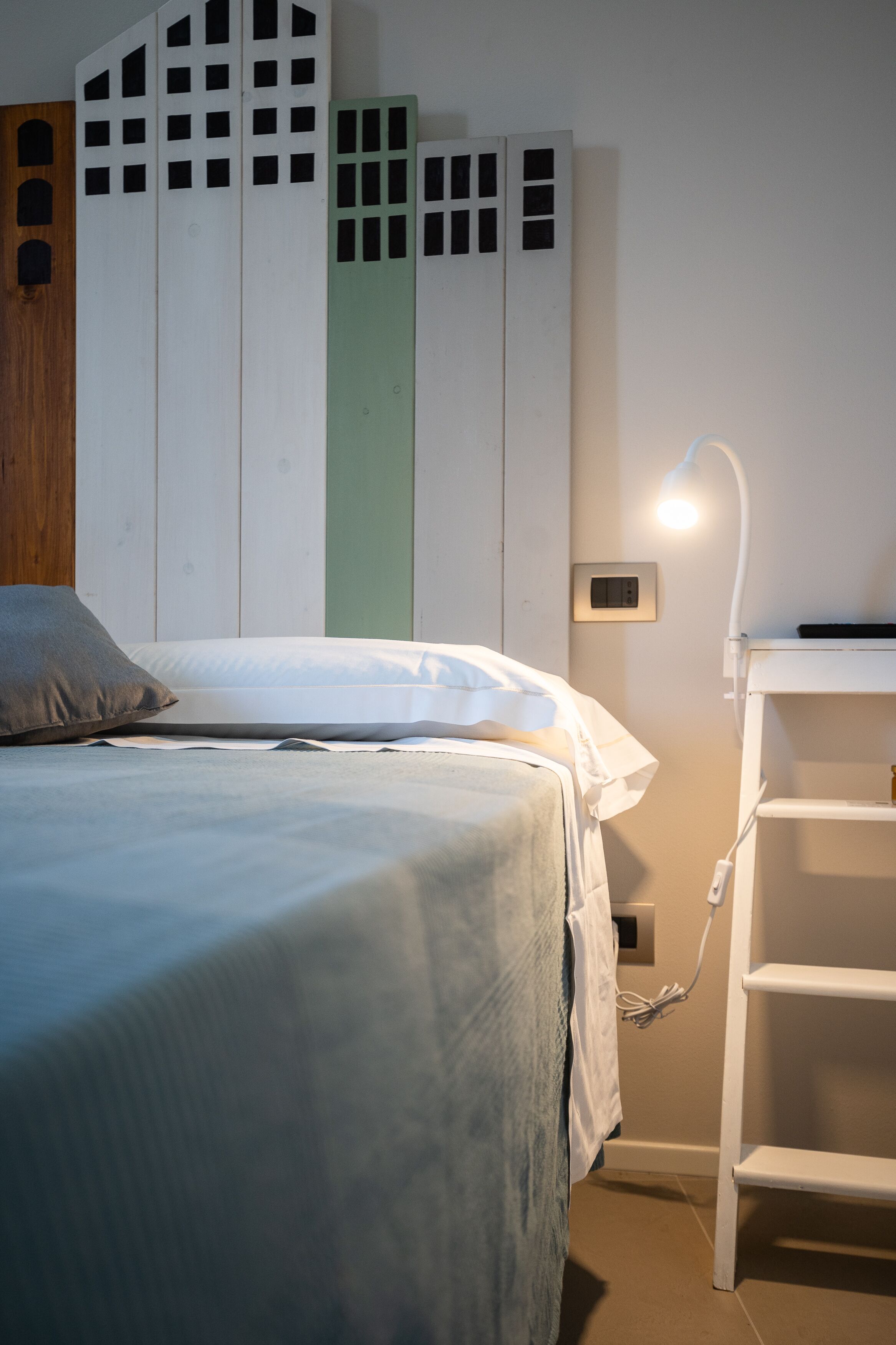 Una cama bien hecha con un almohadón gris, una lámpara de noche y una mesita de noche blanca con toma de corriente.