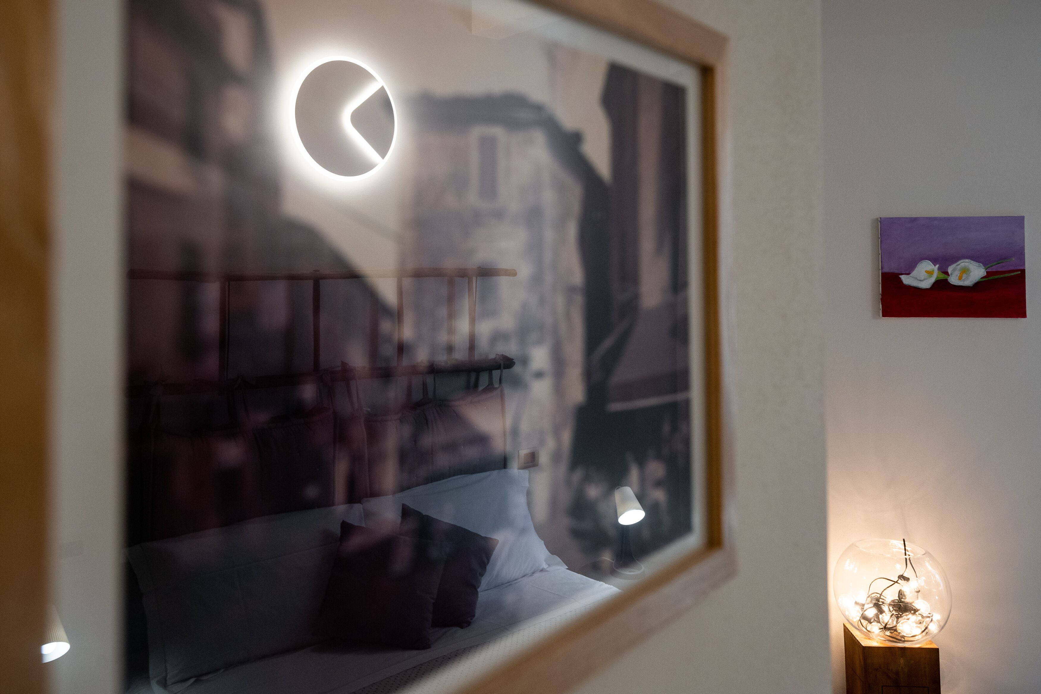 Una stanza con un letto, una finestra che offre vista sugli edifici, una lampada fissata al muro e un tavolino con una lampada.