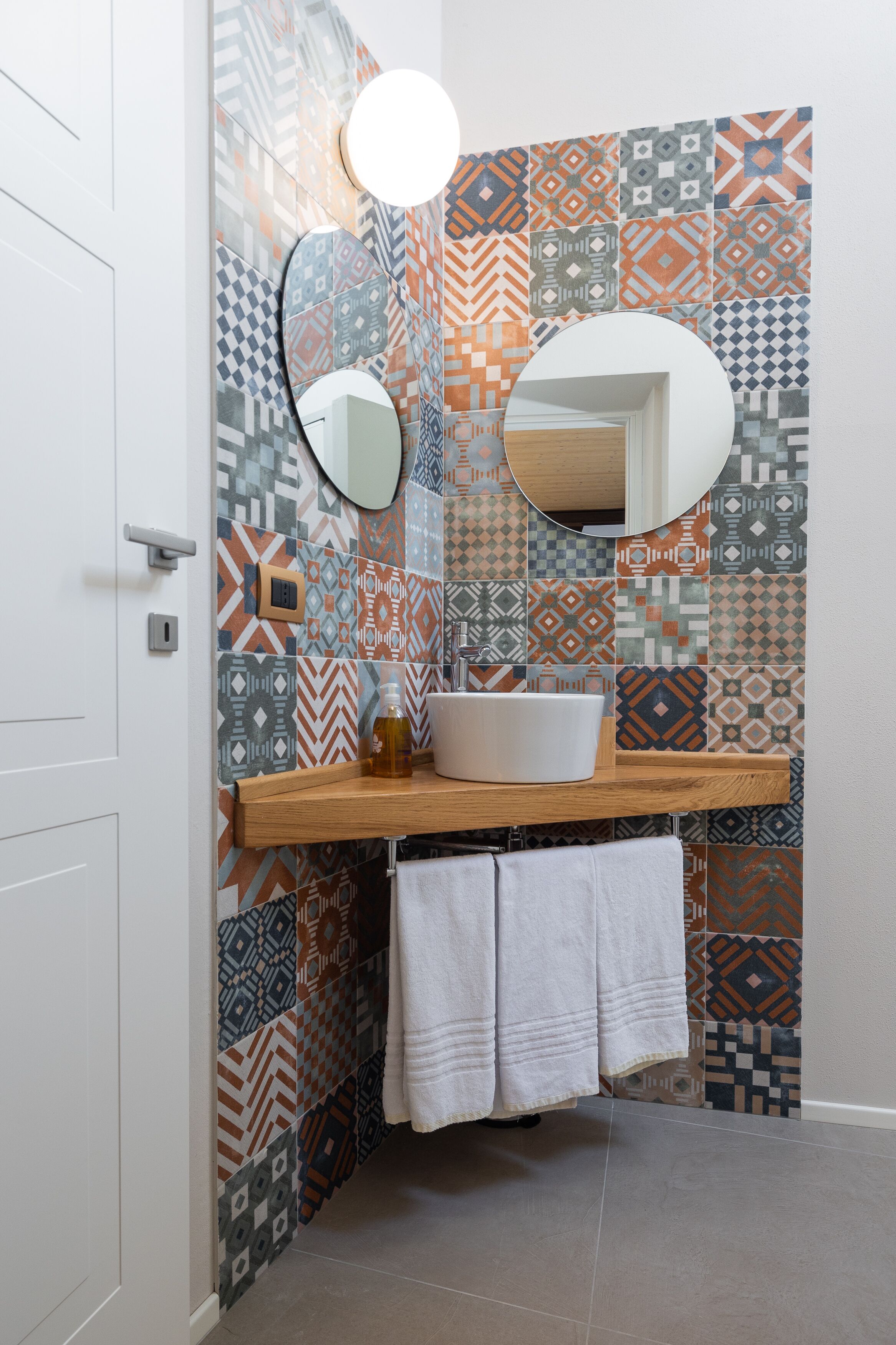 Un baño con azulejos estampados, un lavamanos blanco, dos espejos y un perchero para toallas.