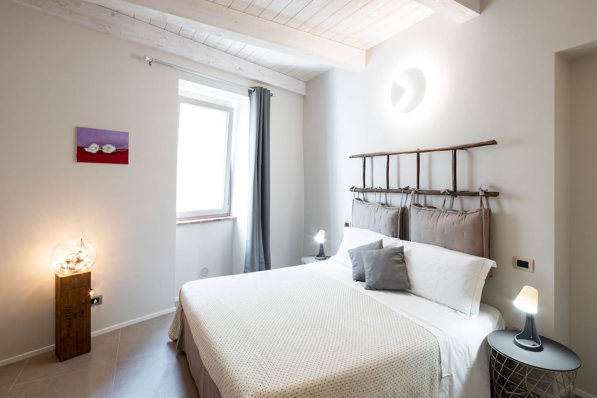 A bedroom with a bed, bedside lamps, a window with curtains, a wall-mounted light fixture, and a painting.