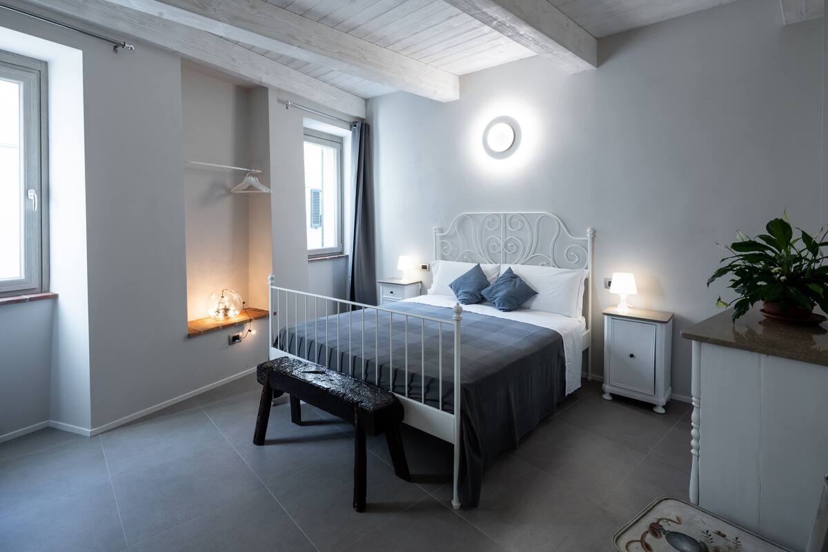 A bedroom with a bed, bedside tables, a bench, a potted plant, and a window.
