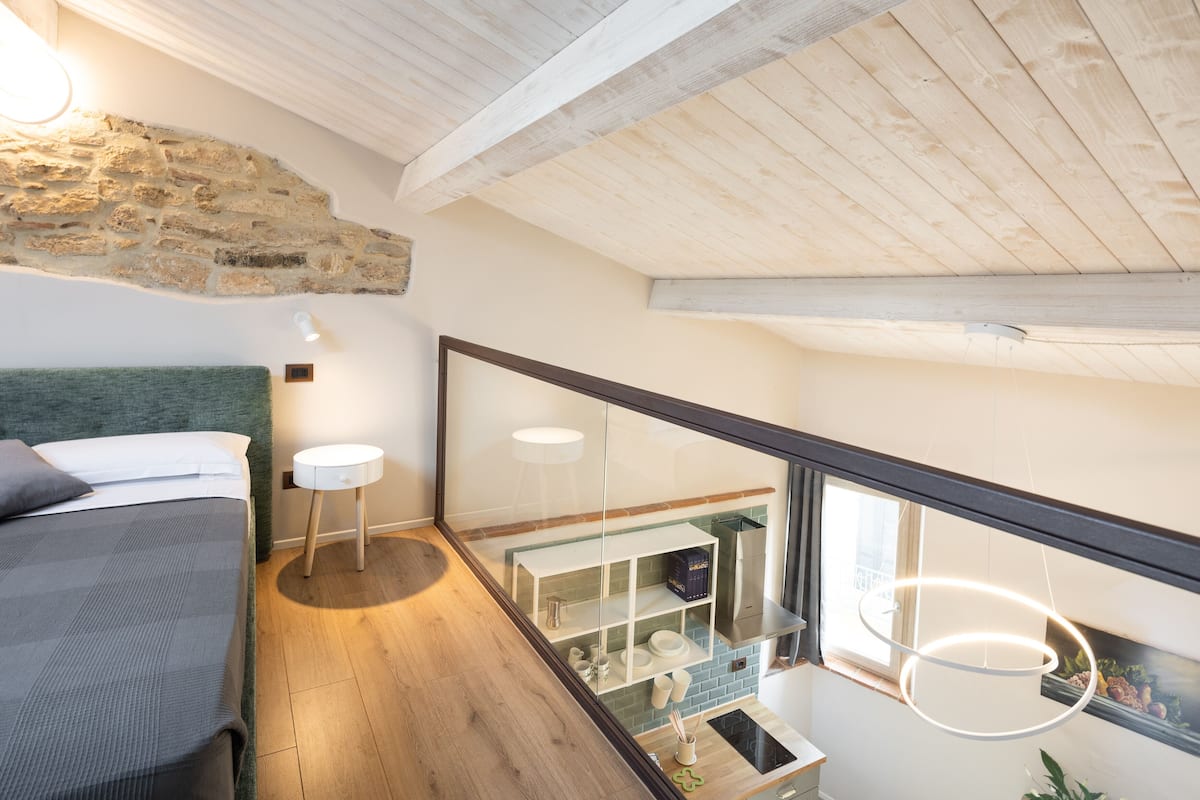 A bedroom with a bed, bedside table, and a view of a kitchen area through a glass partition.