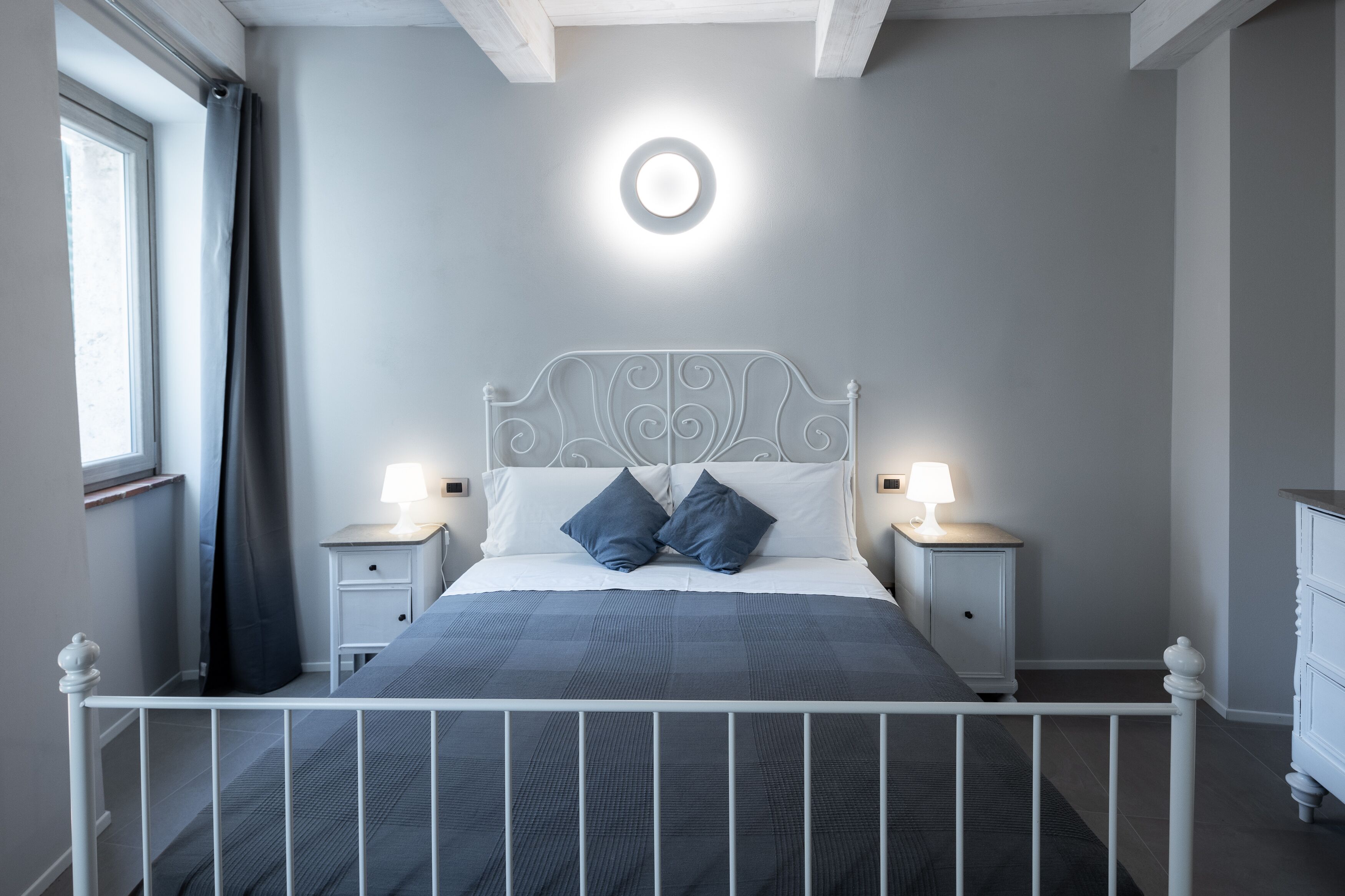 A bedroom with a bed, two bedside tables with lamps, a circular ceiling light, and a window with curtains.