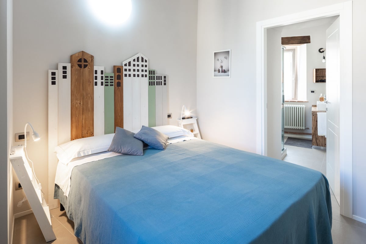 A bedroom with a bed, a nightstand, a wall-mounted lamp, and a door leading to a bathroom.