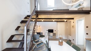 Un interior moderno con una escalera que conduce a un altillo, una mesa de comedor con sillas y un televisor al fondo.