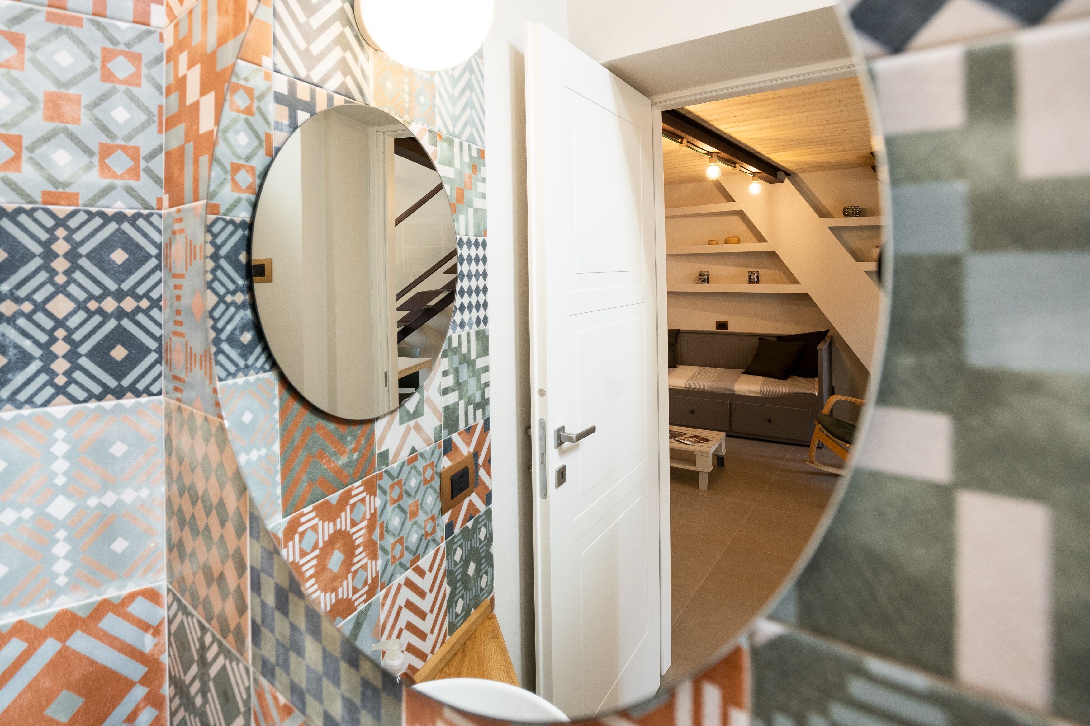 Ein modernes Badezimmer mit gemusterten Fliesen, einem runden Spiegel und einer weißen Tür, die in ein Schlafzimmer mit Bett und Nachttisch führt.