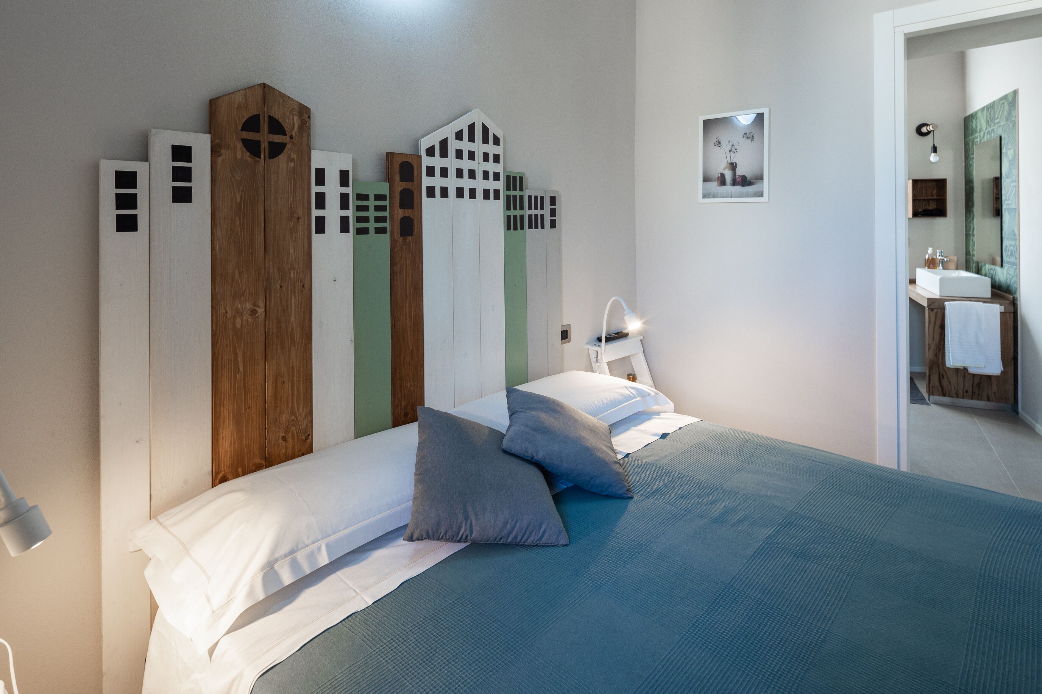Une chambre à coucher avec un lit, des oreillers, une table de chevet, une lampe fixée au mur et un tableau encadré.
