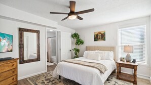 Une chambre à coucher comprenant un lit, un ventilateur de plafond, une table de chevet, un téléviseur, un miroir et une porte donnant sur une autre pièce.