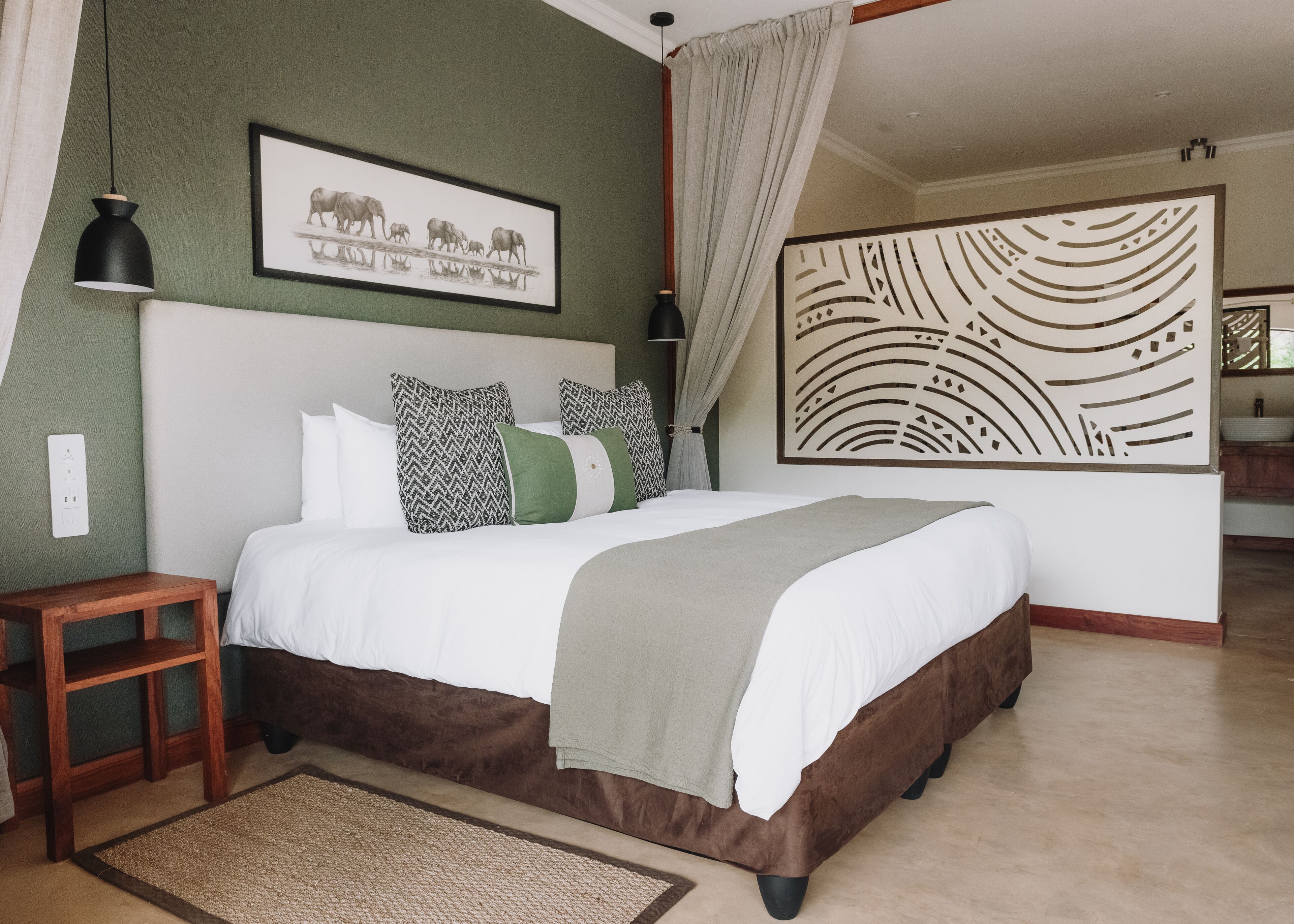 Quarto com cama grande, mesa de cabeceira, criado-mudo e um quadro na parede, além de um painel decorativo.