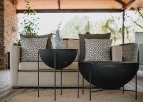 Un área de estar exterior moderna con un sofá y dos mesas de metal negro, un jarrón decorativo y cojines estampados.