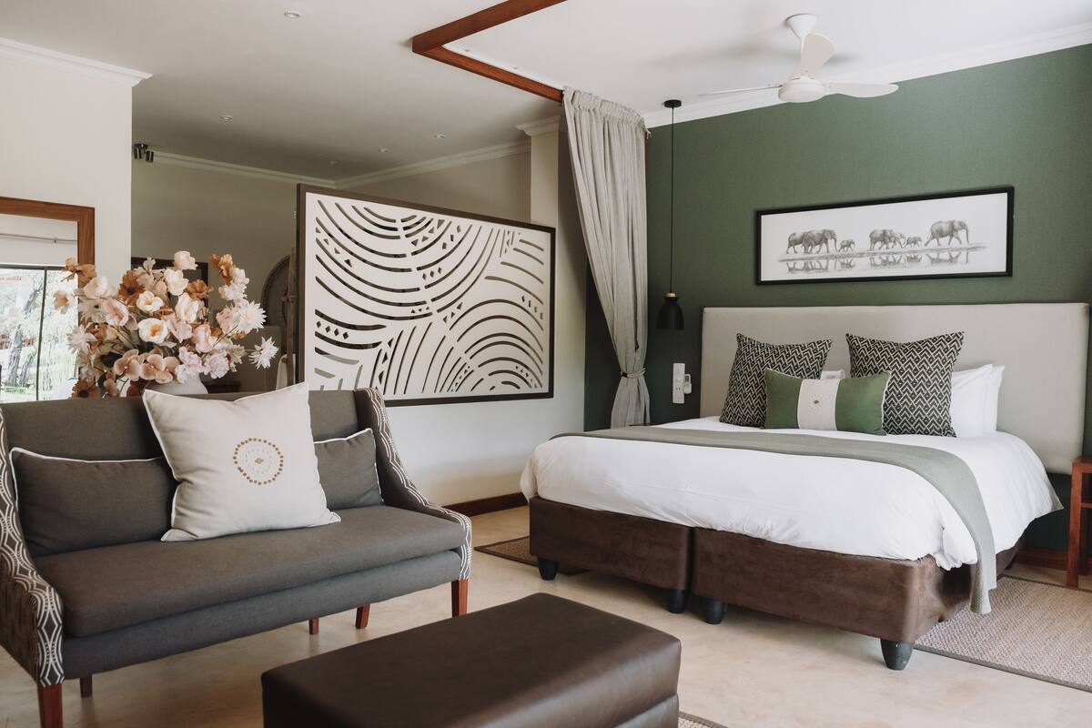 A bedroom with a bed, a sofa, and an ottoman. There is a large wall art piece above the bed and a framed picture on the wall.