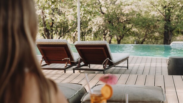 Une personne est assise à une table avec deux boissons, et elle a devant elle une piscine entourée de chaises longues et d’ombrelles.