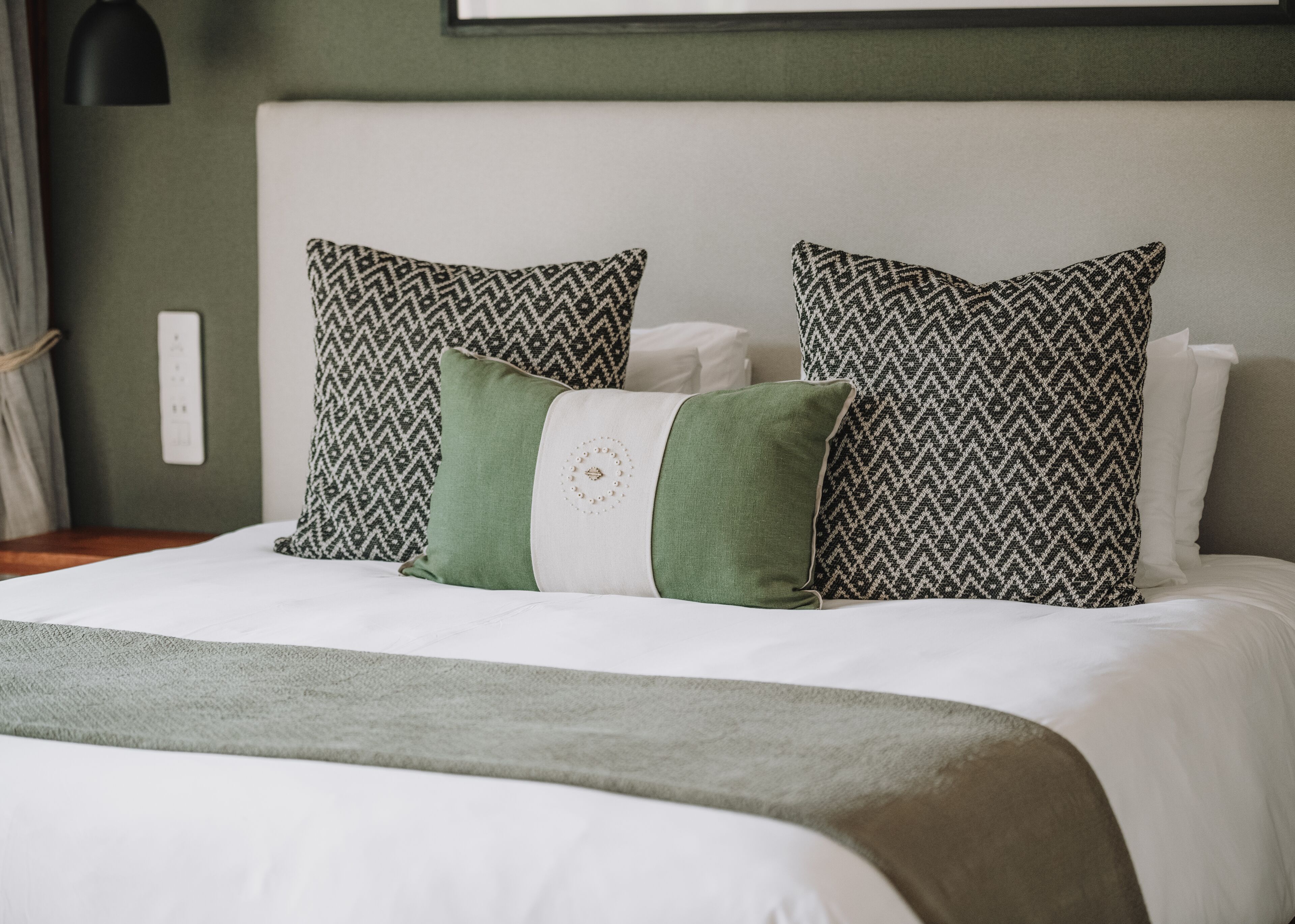 A neatly made bed with patterned pillows, a green throw blanket, and a white headboard.