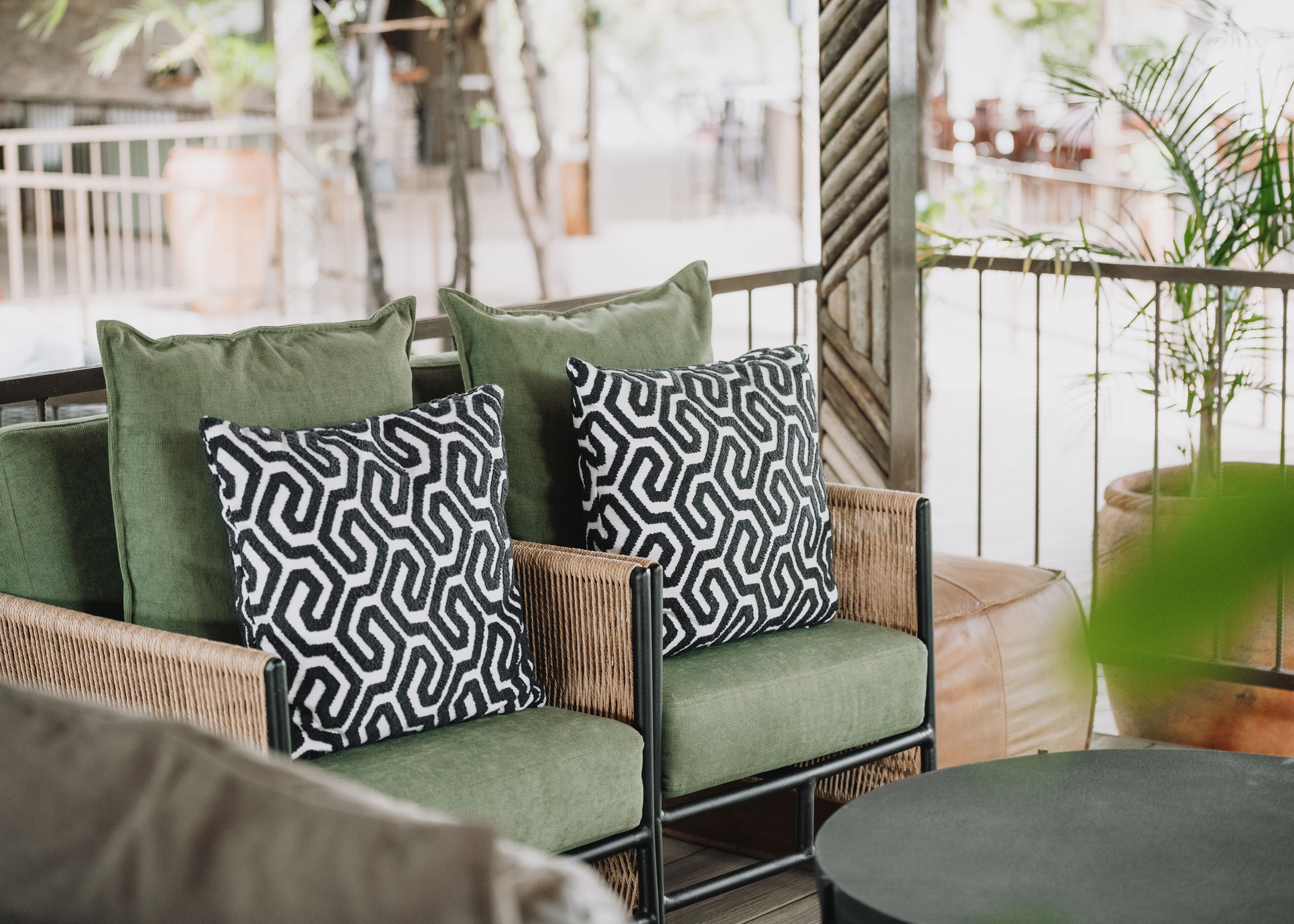 A wicker chair with green cushions and patterned pillows, a round table, and a potted plant.