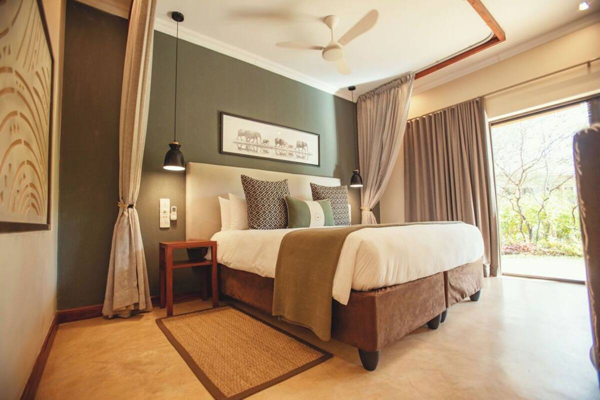 A bedroom with a large bed, a ceiling fan, a nightstand, and a view of the outdoors through a large window.