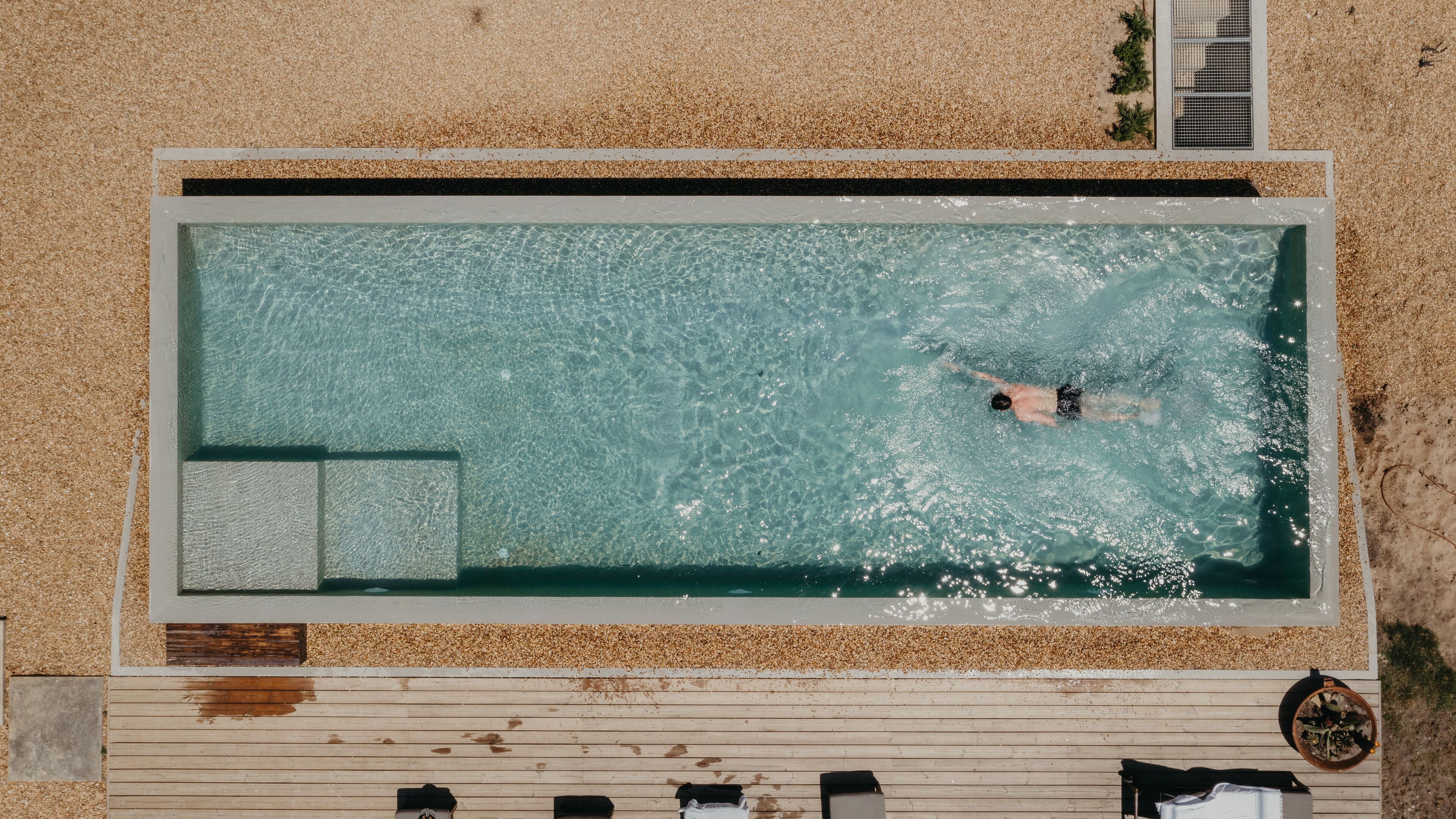 Vue aérienne d’une piscine rectangulaire à l’eau claire, entourée de galets et d’une terrasse en bois.