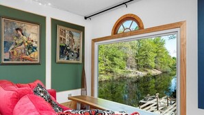A room with a large window overlooking a lake, a red sofa, and two framed paintings on the wall.