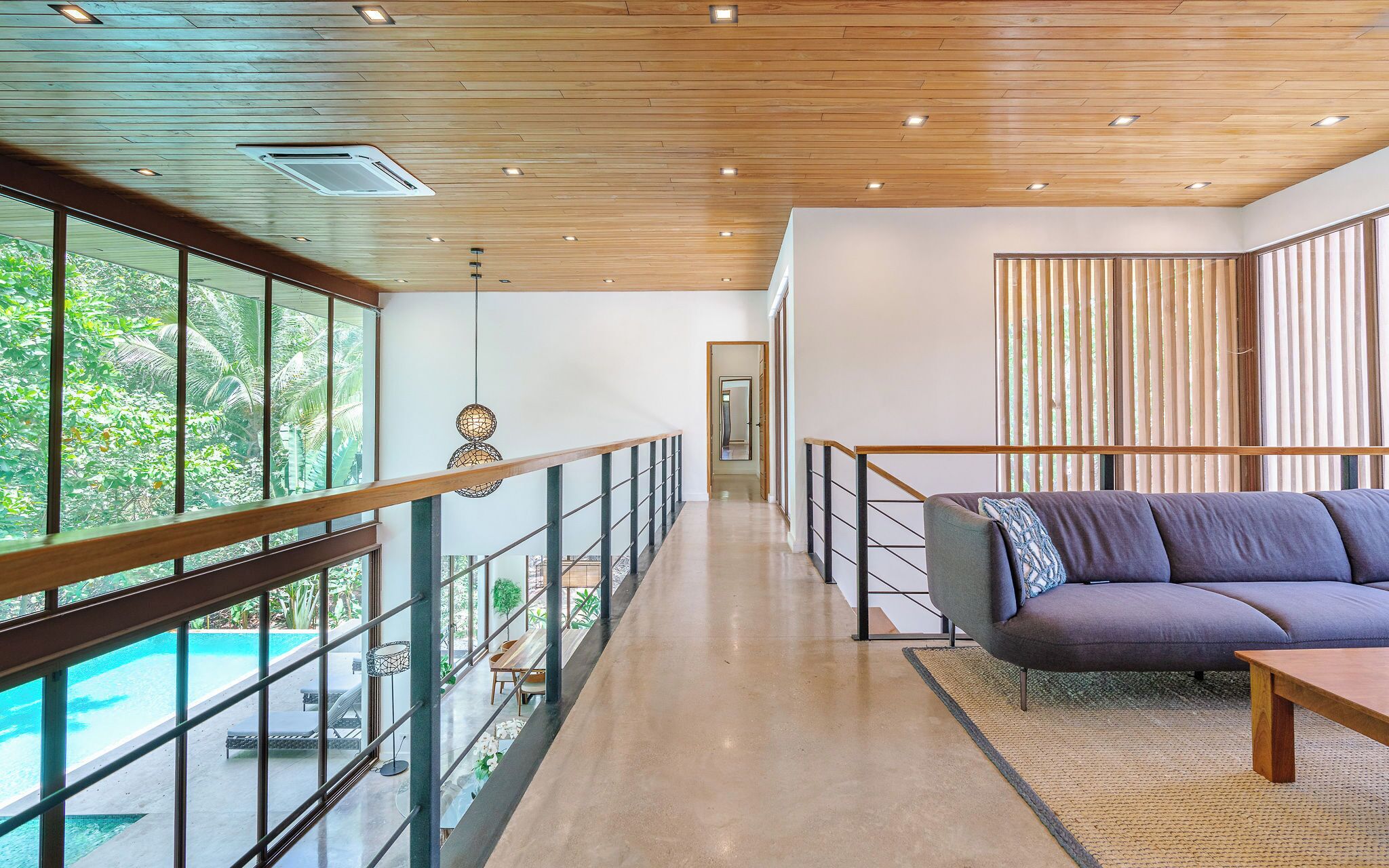 A modern interior with a glass balcony overlooking a pool, a wooden ceiling, and a sofa with a patterned throw.