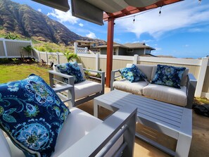 An outdoor patio with white furniture and blue patterned cushions, set against a backdrop of a mountain and clear sky. - Brand New 4BHK Home! Ocean and Mountain View! (Waianae)