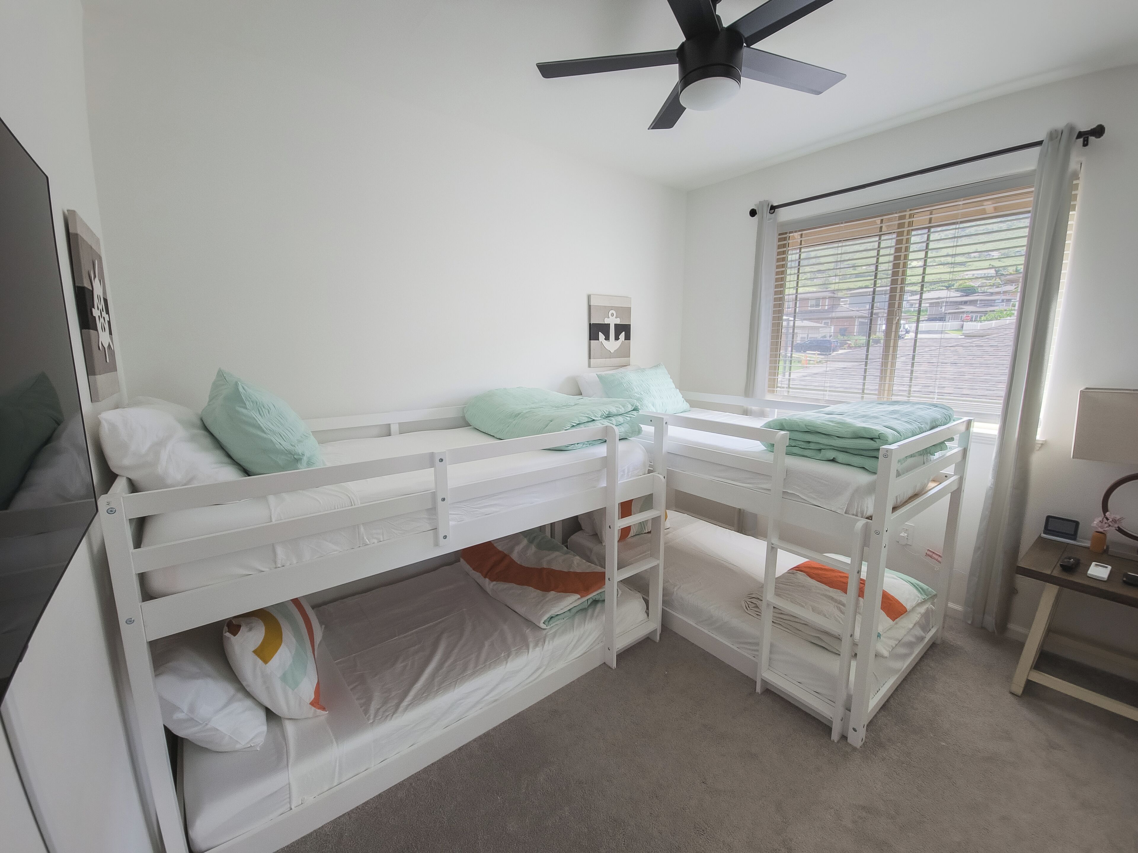 A bunk bed room with white bedding, a ceiling fan, a window with blinds, and a small table with a lamp.