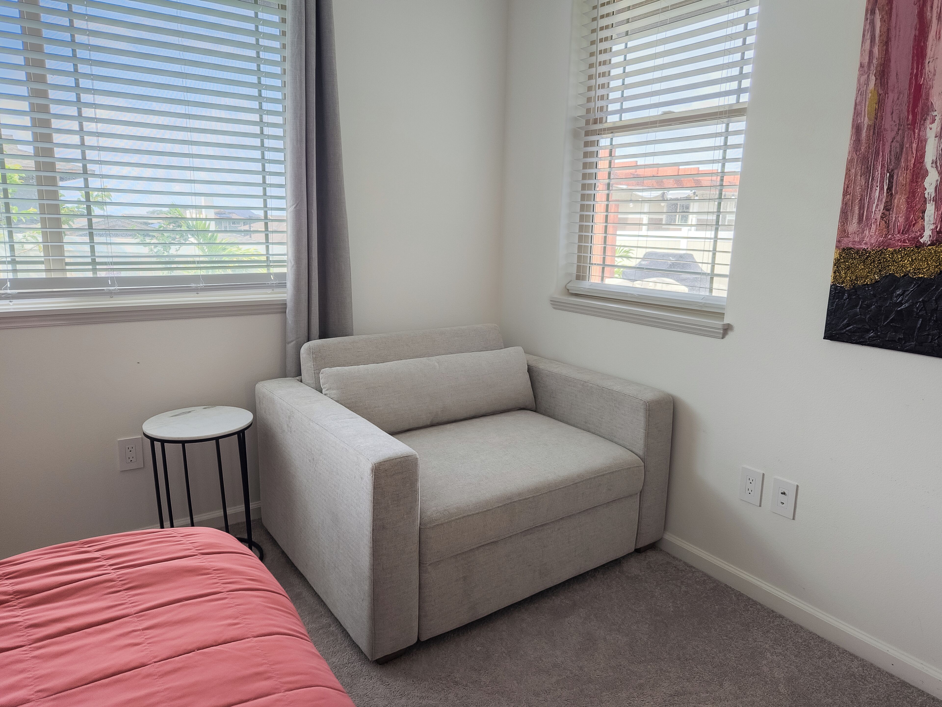 A room with a grey sofa, a small round table, and a bed with a pink cover.