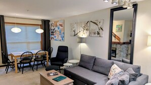 Ein modernes Wohnzimmer mit grauem Sofa, einem Holztisch, einem schwarzen Sessel und einem Essbereich mit Holztisch und Stühlen. An den Wänden befinden sich Kunstwerke und ein Spiegel.