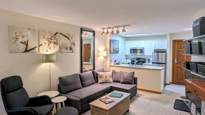 Ein modernes Wohnzimmer mit einer Eckcouch, einem Couchtisch und einem schwarzen Sessel. Im Hintergrund ist eine Küche mit weißen Schränken und Edelstahlgeräten zu sehen.