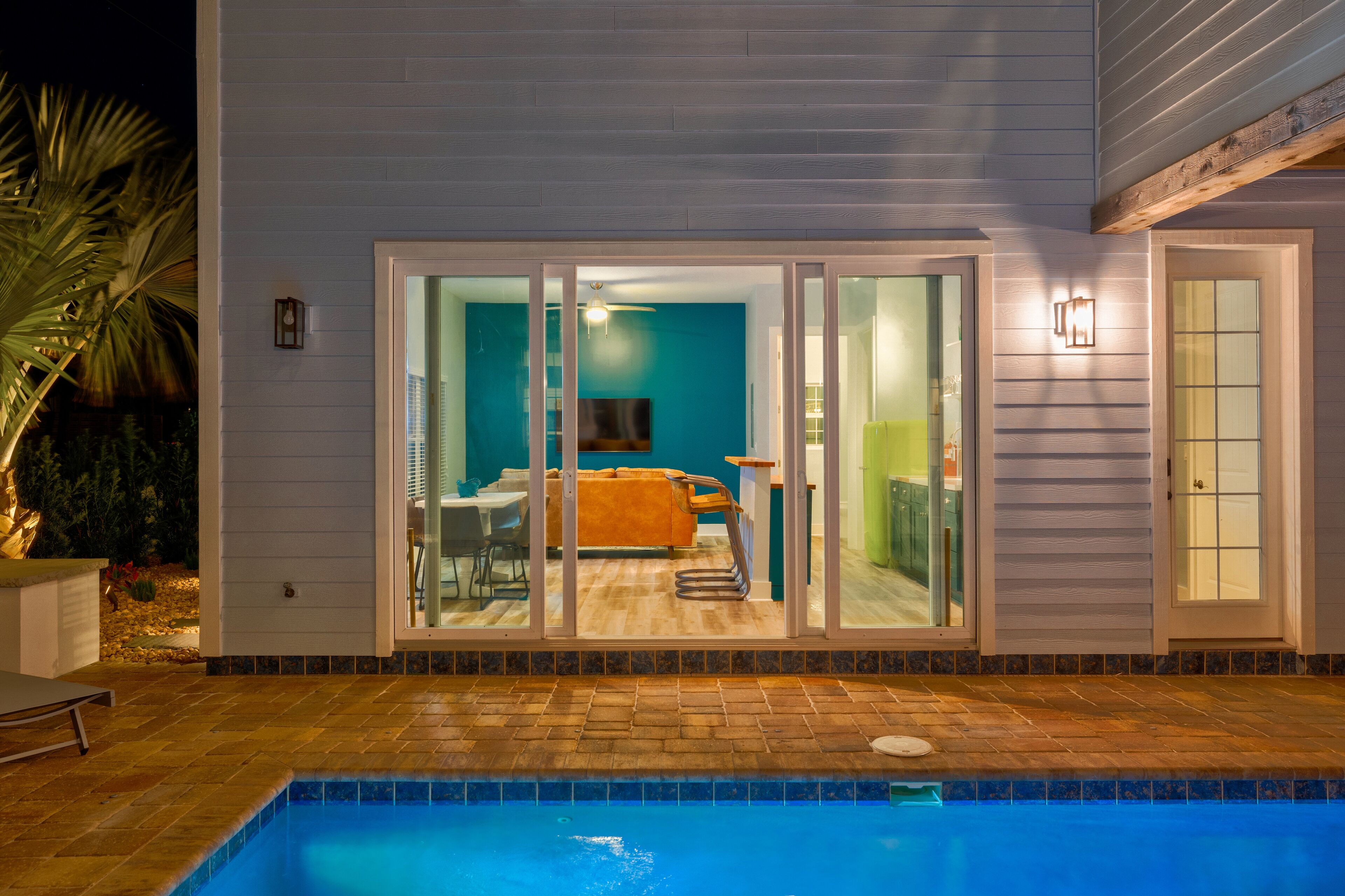 A swimming pool with a tiled edge, a sliding glass door leading to a room with a sofa and a television, and a small outdoor seating area with a chair.