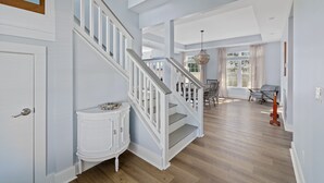 A spacious hallway with a wooden staircase, a white console table, and a dining area with a table and chairs. - Oversized home w/ Heated Pool - mins from beach! (St. Augustine)