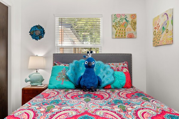 Un dormitorio con una colcha colorida, un cojín con motivos de pavo real, un adorno de pared y una ventana con persianas.