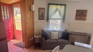 Ein gemütliches Wohnzimmer mit einer Couch, einem kleinen Tisch, einem Sessel, einem Fenster mit Vorhang und einer Tür, die nach draußen führt.