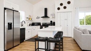 A modern kitchen with a stainless steel refrigerator, a microwave, and a coffee machine. There is a white island with bar stools and a white sofa in the living area. - Modern Luxury 5BR 4BA with Heated Pool and EV Charge (Branson)