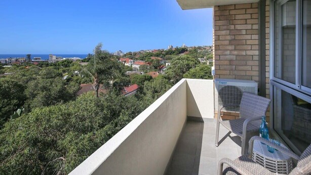 Un balcón con mobiliario de mimbre, vistas a un pueblo costero y cielo despejado.