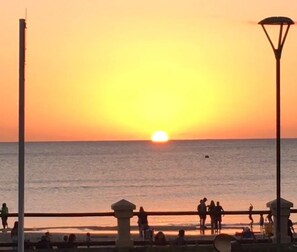 Un atardecer sobre una playa con gente caminando por la orilla y una barandilla al frente.
