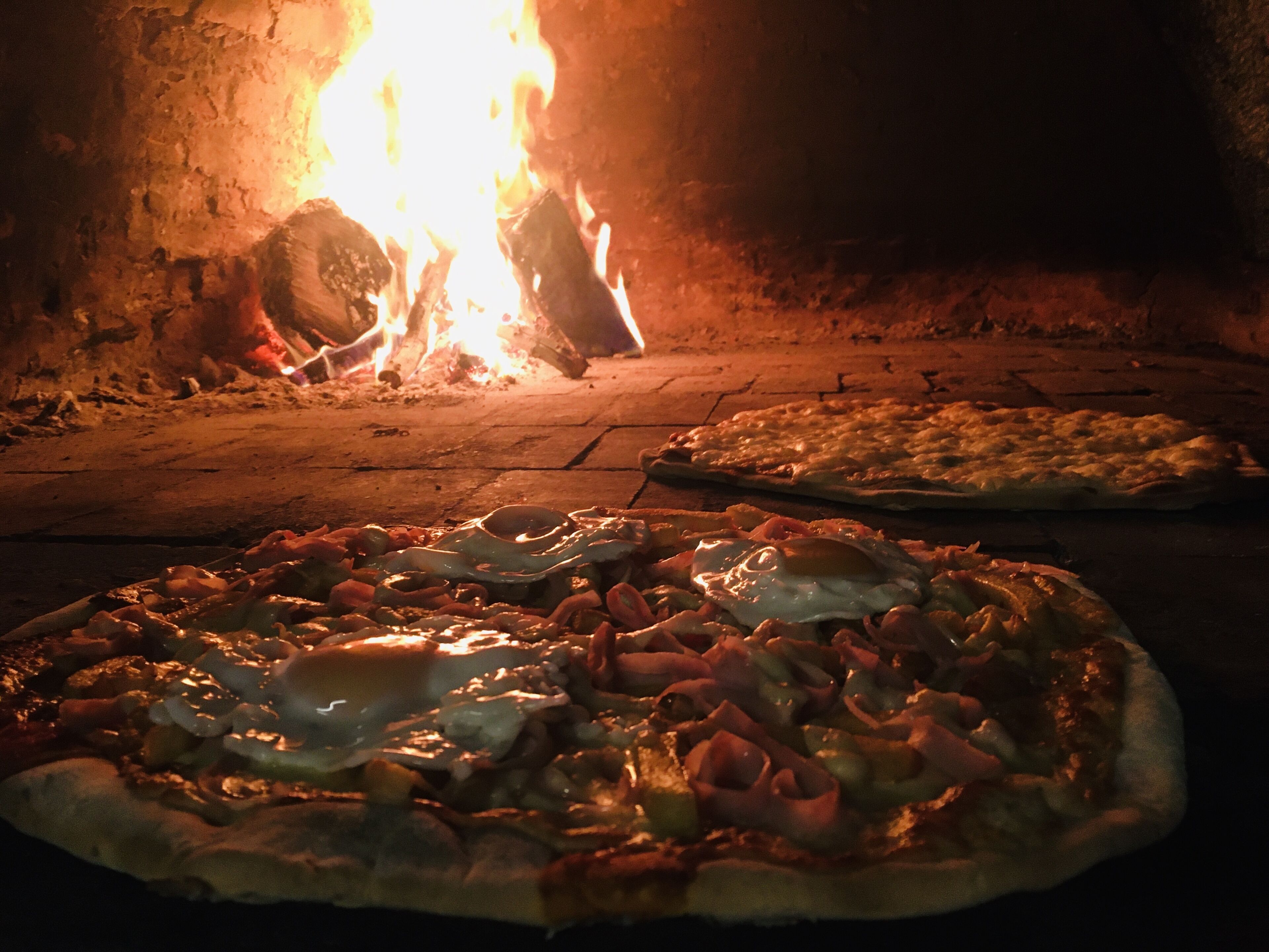 Due pizze in cottura in un forno a legna, con fiamme visibili sullo sfondo.