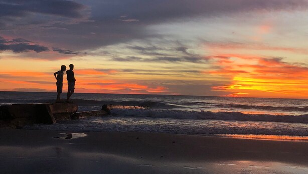 A beach scene with two silhouetted figures standing on a concrete barrier, overlooking the ocean during sunset. - Hotel Alcázar  (Piriapolis)