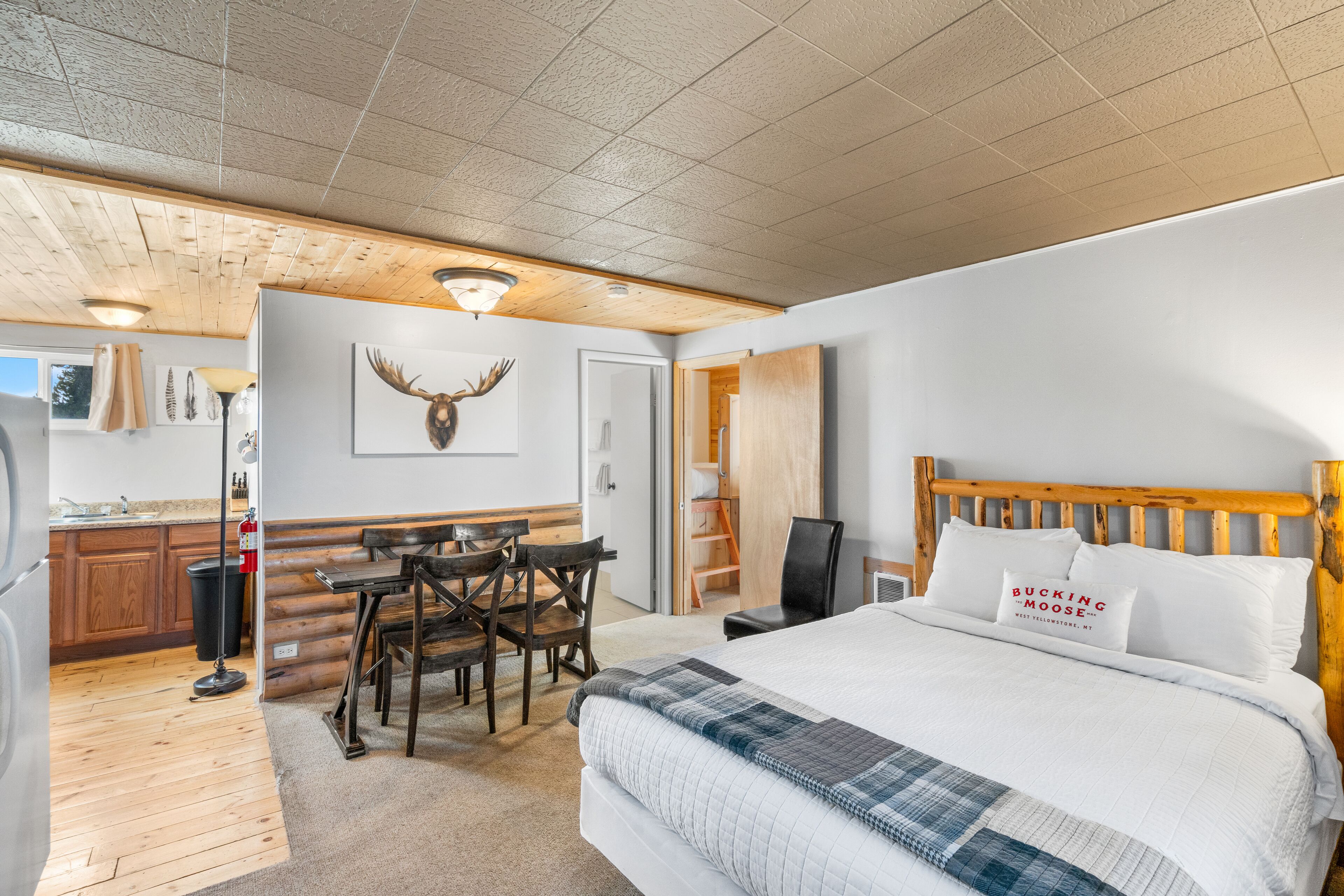 A bedroom with a large bed, wooden headboard, a dining area with a table and chairs, a kitchen area, and a closet.