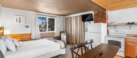 Quarto compacto com cama, mesa de jantar de madeira, cozinha equipada com fogão e geladeira, e janela com cortinas.
