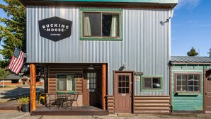 A rustic building with a sign reading "Bucking the Moose Cabins." It features a wooden door, a small porch with a table and chairs, and a window with curtains. - Experience comfort at The Bucking Moose (#02) The Loft, just 1 minute from Yellowstone! (West Yellowstone)