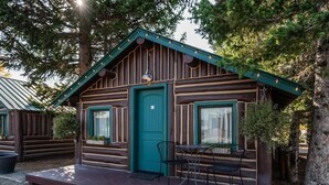 A rustic cabin with a green door, a small porch with a table and chairs, and a window with a curtain. - Cozy Bucking Moose, Moose Cabin with 2 Queens, just a minute from Yellowstone! (West Yellowstone)