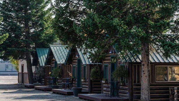 A row of log cabins with green roofs, outdoor seating, and trees in the background. - Cozy Bucking Moose, Moose Cabin with 2 Queens, just a minute from Yellowstone! (West Yellowstone)