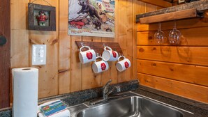 A wooden cabin interior with a kitchen area, including a sink, hanging mugs, and a poster on the wall. - Cozy Bucking Moose, Moose Cabin with 2 Queens, just a minute from Yellowstone! (West Yellowstone)