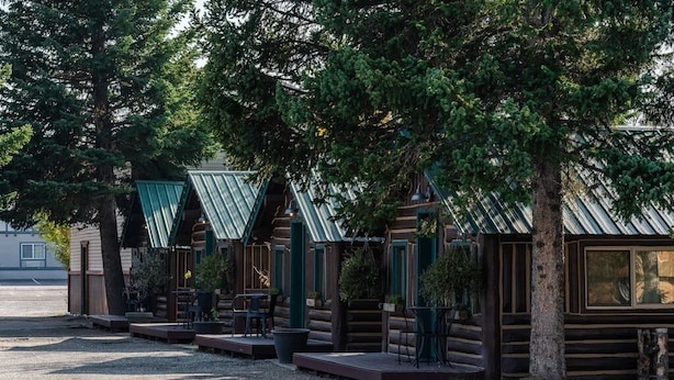 Una hilera de cabañas de madera con techos verdes, mobiliario exterior y árboles al fondo.