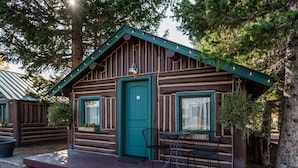 Una cabaña rústica con puerta verde, un porche peque ño con mesa y sillas, y una ventana con cortina.