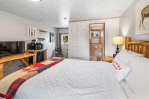 A hotel room with a bed, a desk with a TV and microwave, a chair, and a wooden nightstand with a lamp.