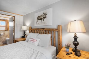 A bedroom with a wooden headboard, a bed with white bedding, a moose artwork on the wall, a bedside lamp, and a small table with a plant and a remote control.