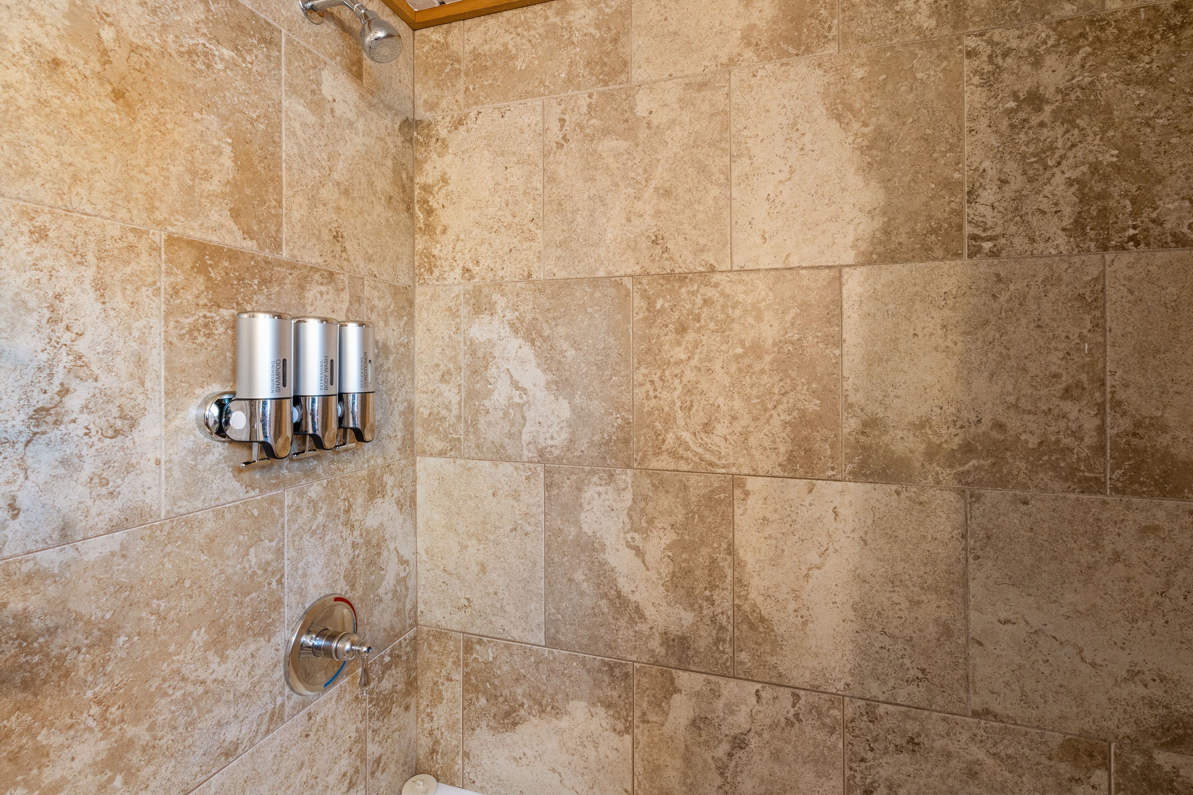 Un baño con azulejos color beige, una alcachofa de ducha y tres dispensadores metálicos en la pared.